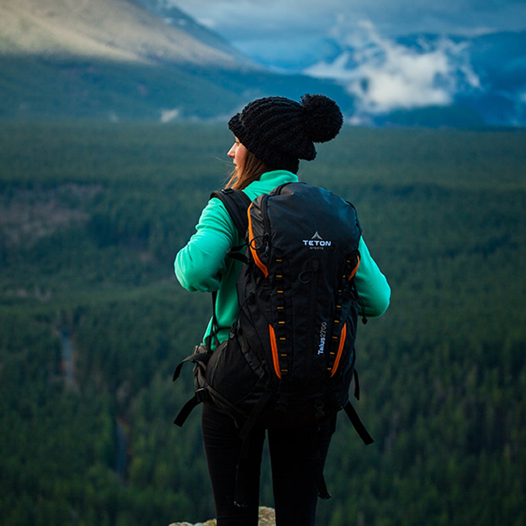 Woman stands looking over cliff with backpack.