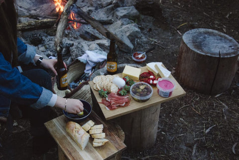 A pair of hands making a charcuterie board at a campsite by a campfire.