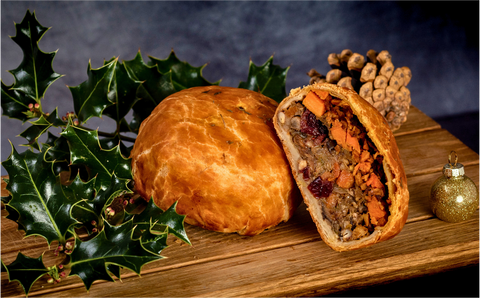Vegan wellington with a cut open wellington resting against it, showing a filling of lentils, vegetables and vranberries. The wellington is on a wooden board next to sprigs of holly, a pine cone and a gold bauble.