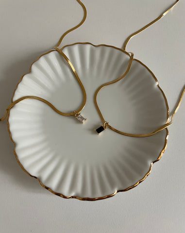 Coupelle en porcelaine blanche de forme ronde sur laquelle sont posés deux colliers en acier inoxydable doré avec une chaine serpentine et un pendentif en zircon couleur diamant pour l'un et de couleur noire pour l'autre