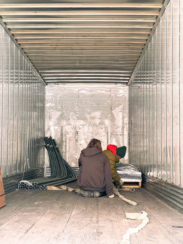 Two men kneeling on the floor working on unloading a palette of materials from the back of a tractor trailer.