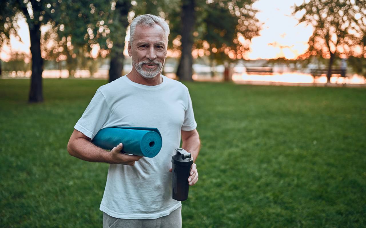 Senior man doing sport outdoors while holding a bottle of bone broth and a yoga mat