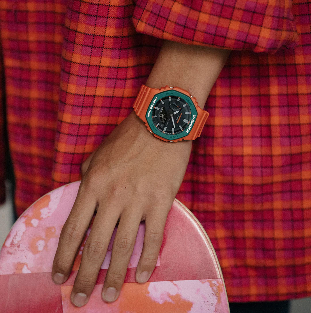 G-Shock Skater Flavour GA211-SC-4A. Orange watch worn on wrist with hand on skateboard. 