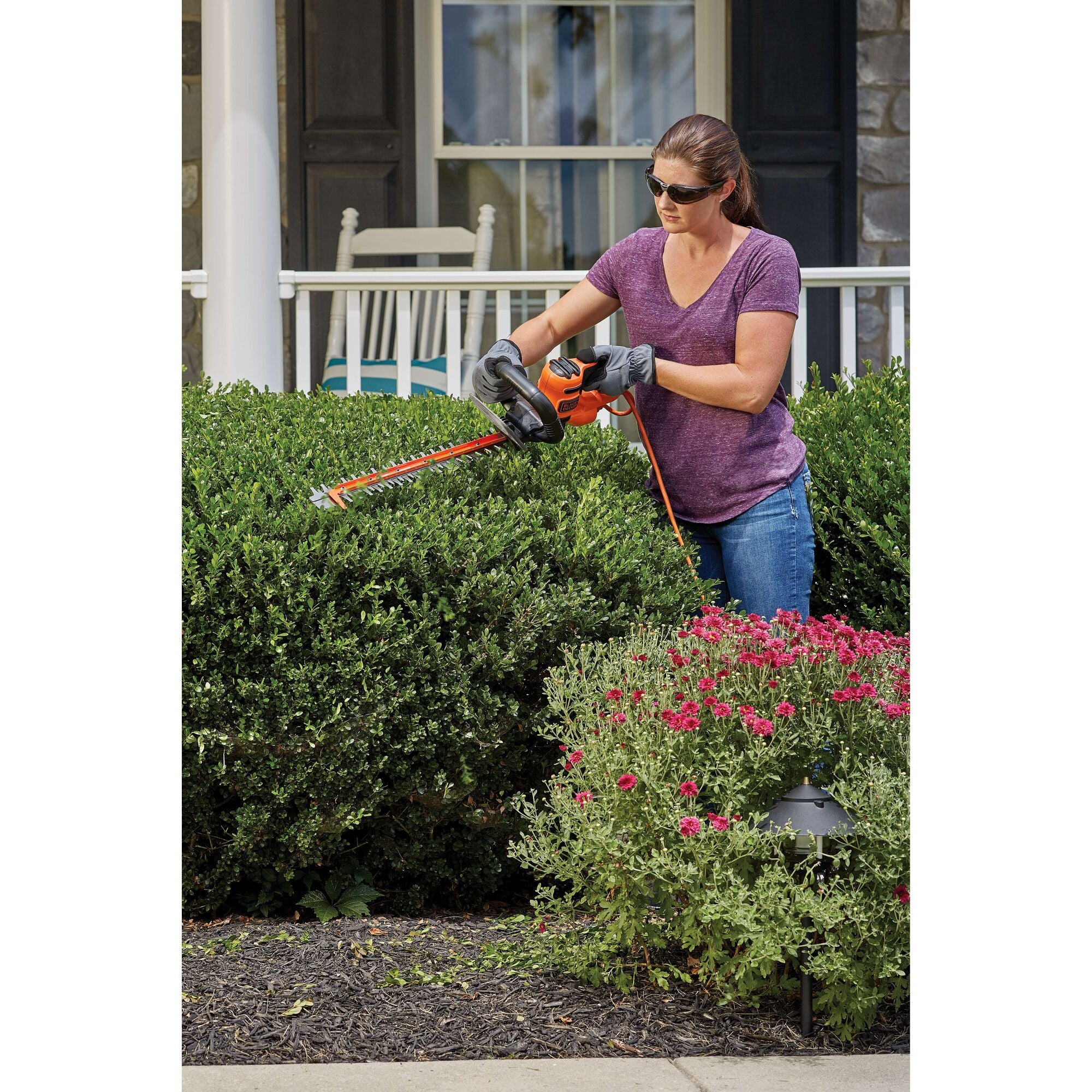 A woman is trimming bushes using a BLACK+DECKER Hedge Trimmer.