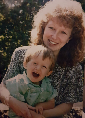 Three year old boy with red hair and fair skin, sits on his Mum's lap with a wide mouth laughing. Mum with curly red hair and fair skin smiles and wraps her arms around him