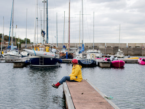 MJ sits on the end of a marina pontoon. there are sail boats in the background and she is dressed in a bright red hat and boots, a yellow oilskin jacket and jeans.