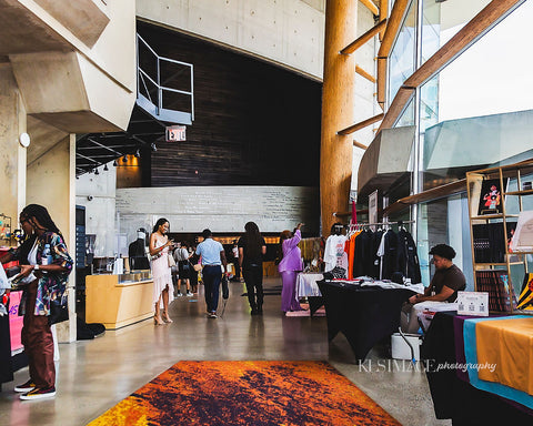 A photograph of the lobby of Arena Stage during a BIPOC event called "Melanincon". There are various people setting up their vending stations, and Donny is in the lower right hand corner with a brown short sleeve shirt. To the right of Donny is a clothing rack filled with 17th & Granville merchandise.