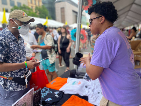 An image of 17th & Granville owner Donny in the purple choices t-shirt featuring a graphic on the back of the shirt of a man holding a deck of cards that say life coach, generational wealth, support system and therapist. Donny also has a short curly afro with sunglasses on while talking with a masculine customer at the 2023 Capital Pride Festival in DC. The conversation is about 17th & Granville being inclusive, gender neutral and a brand for everyone. We advocate for and empower BIPOC and LGBTQ+ communities. In front of Donny there is a table with various 17th & Granville products including the Love You More Summer Edition T-shirt, Black Queer Excellence T-shirt and Orange 17th & Granville Signature Shorts. In the background there are various patrons enjoying the festival.