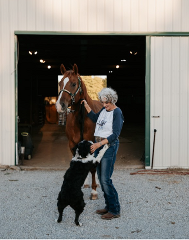 A dog jumping into a woman's arms while the woman and a horse stand in front of a horse stable