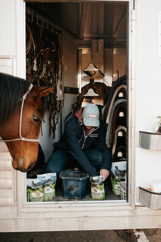 A woman kneeling over and scooping out herbs to an eager-looking horse nearby
