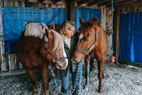 A woman between two horses, giving both horses a hug inside a horse stable