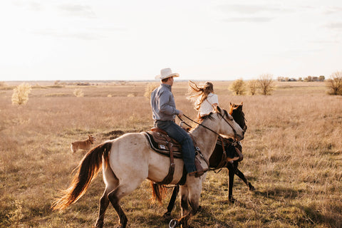 A man and woman riding a horse in a field with a dog trailing behind