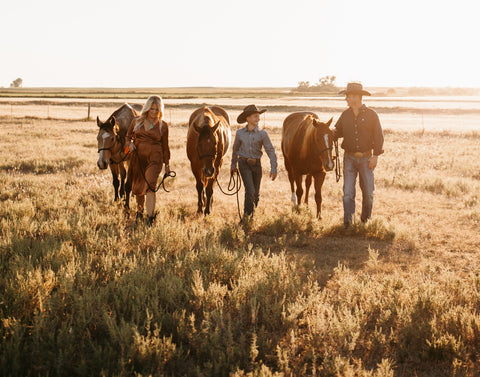 A man, woman, and young boy walking in a shadow-laden field of grass with horses