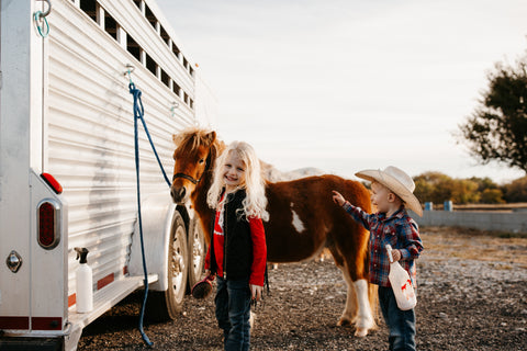 Two toddlers with a water bottle in their hand smiling, and a small horse in the background near a trailer