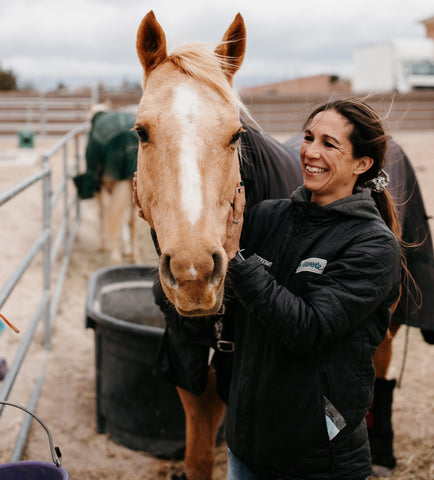 A woman helping smooth the coat of a brown horse, with the horse staring at the camera and the woman smiling