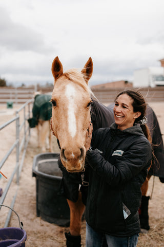 A woman smiling and looking at her horse with her hands on her horse's mane; the horse is looking at the camera