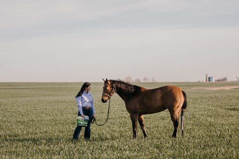A woman in a field of grass smiling and looking her horse in the eyes