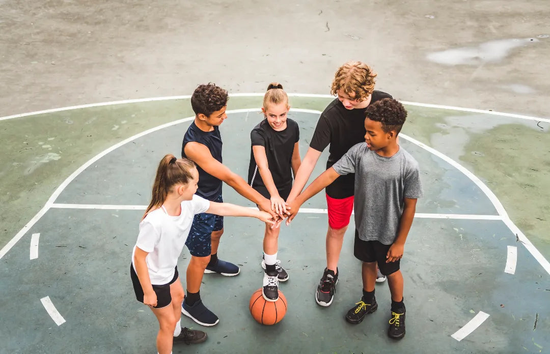 Group of children putting hands together over a basketball on an outdoor court.