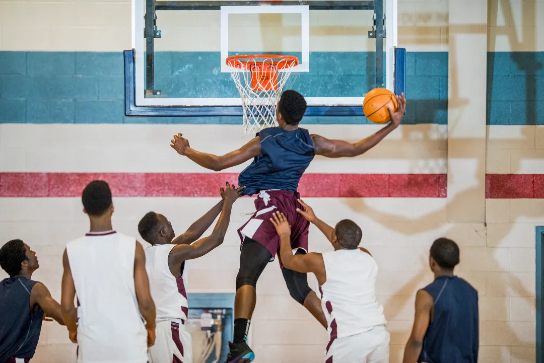 A basketball player in a dark jersey jumps to score amidst defenders in a gym.