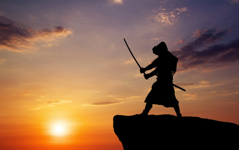 Silhouette of samurai on cliff side wielding katana in an attack stance with a sunset behind