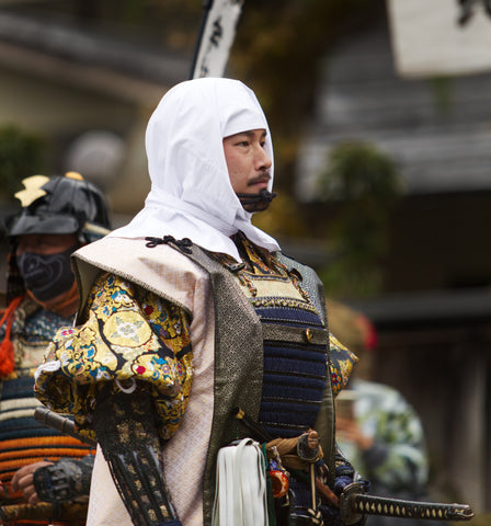 Samurai standing with white covering looking right at an outdoor setting of a dojo