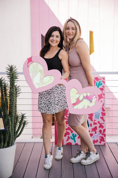 Two smiling women pose with a pair of giant prop heart shaped sunglasses made out of cardboard. 