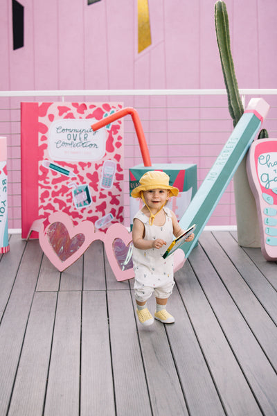 A small baby in a yellow hat stands in front of an art installation of giant cardboard novelty props