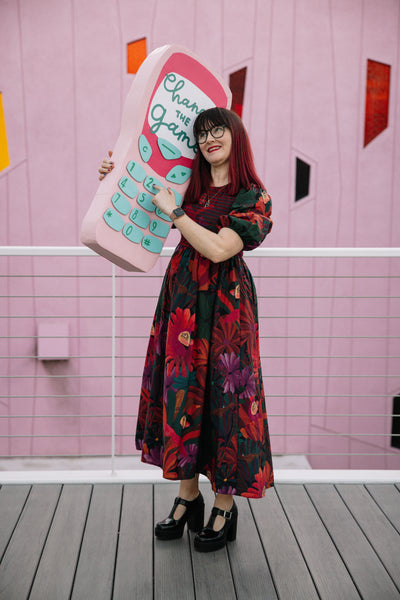 A woman with red hair and a floral dress stands on a balcony holding an oversized Nokia3210 mobile phone prop made from cardboard