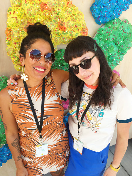 Nikki and Kit, two creative women, stand in front of a colourful backdrop with their arms around each other.