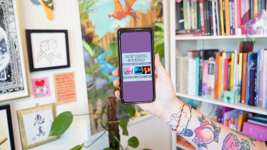 A hand holds up a phone with an Adobe Express template displayed on screen. In the background is a colourful art studio