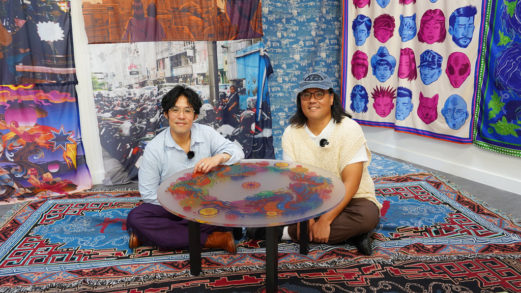 Chris and Andrew, two Asian men, sit on the floor at a round table, surrounded by colourful hanging textiles and rugs