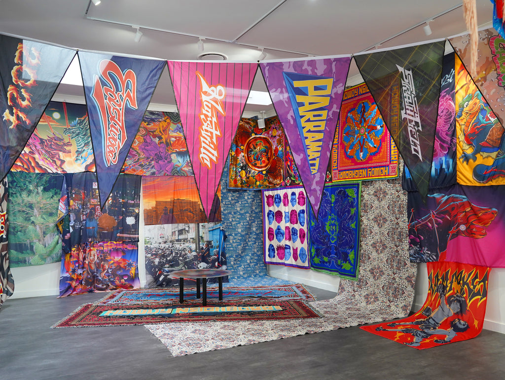 A colourful art installation featuring hanging pennant flags, and scarves. On the floor are vibrant textiles and rugs, and a round decorative table.