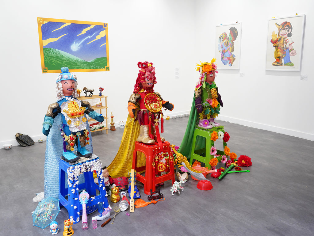 A vibrant and colourful art installation featuring 3 statues surrounded by found objects. In the background, colourful prints hang on the wall.