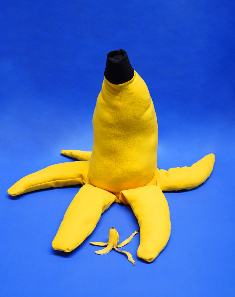 A giant plushie banana prop sits on a blue backdrop. In front of it is a real-sized banana.