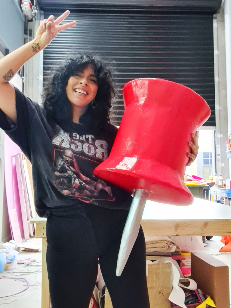 Kitiya, a woman of colour with black hair, a blue sweater and green checkered pants stands in her colourful art studio holding a giant, oversized red papier mâché pin/thumbtack prop, and giving a peace sign