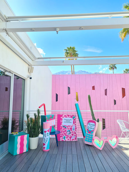 Colourful oversized props sit on a balcony in the sun in front of a pink wall