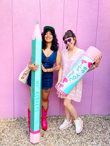 Nikki and Kit, two female designers, stand in front of a pink wall holding a giant handmade novelty pencil and glue stick prop