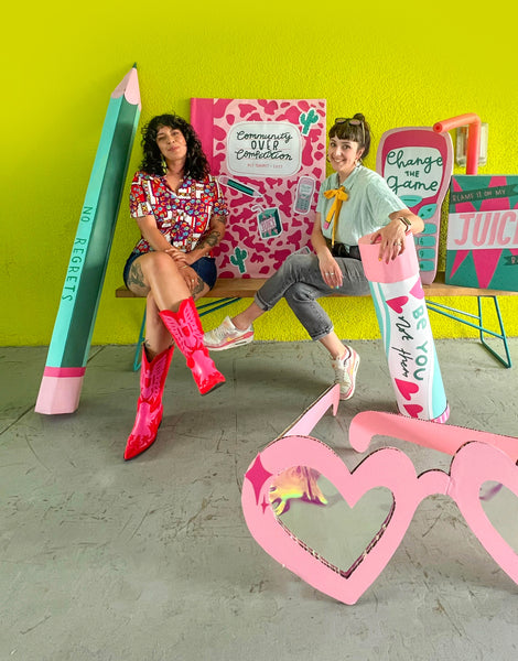 Kit and Nikki, two female designers, sit on a bench in front of a lime green wall, surrounded by handmade oversized novelty cardboard props