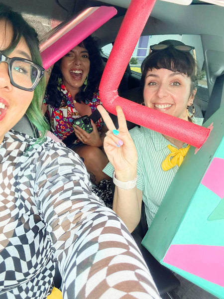 Three laughing women sit in a car that is full of giant novelty cardboard props.