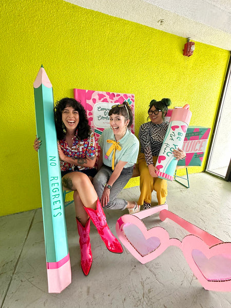 Three smiling women sit on a bench surrounded by giant novelty cardboard props, against a lime green wall.