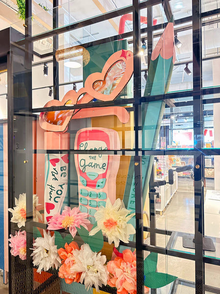 A collection of giant novelty cardboard props is displayed in a shop window surrounded by paper flowers