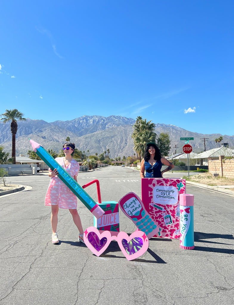Nikki and Kit, two female designers, stand in a street against a desert mountain backdrop, surrounded by giant novelty cardboard props
