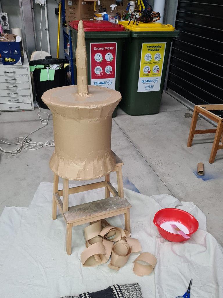 A giant pin/thumbtack prop sits on a stool in an industrial studio space. It is in the process of having papier-mache applied to it and is unpainted.
