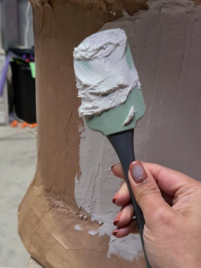 A brown hand with decorated nails holds a silicone spatula up in front of a giant papier-mache pin/thumbtack prop. The spatula is coated with multi-purpose joint compound.