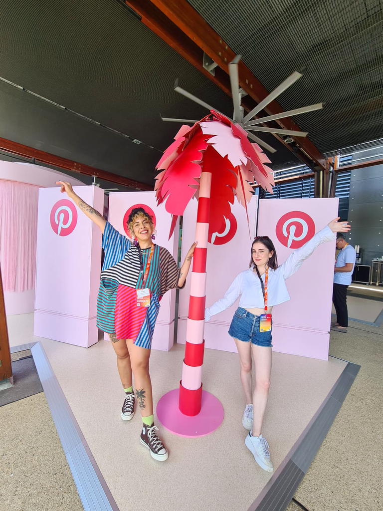 Kitiya and Greta, two creative women, stand in front of a giant papercraft red and pink palm tree that they made, at a Pinterest conference booth.