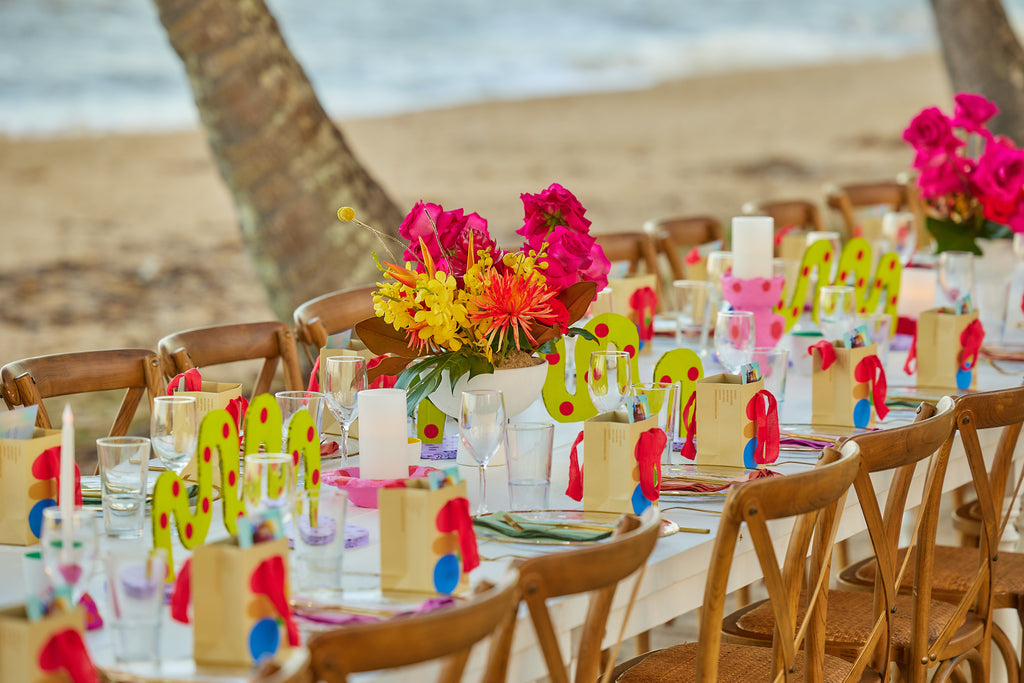 A long dining table on a tropical beach. On the table is a collection of colourful, handmade cardboard decorative table decor pieces.