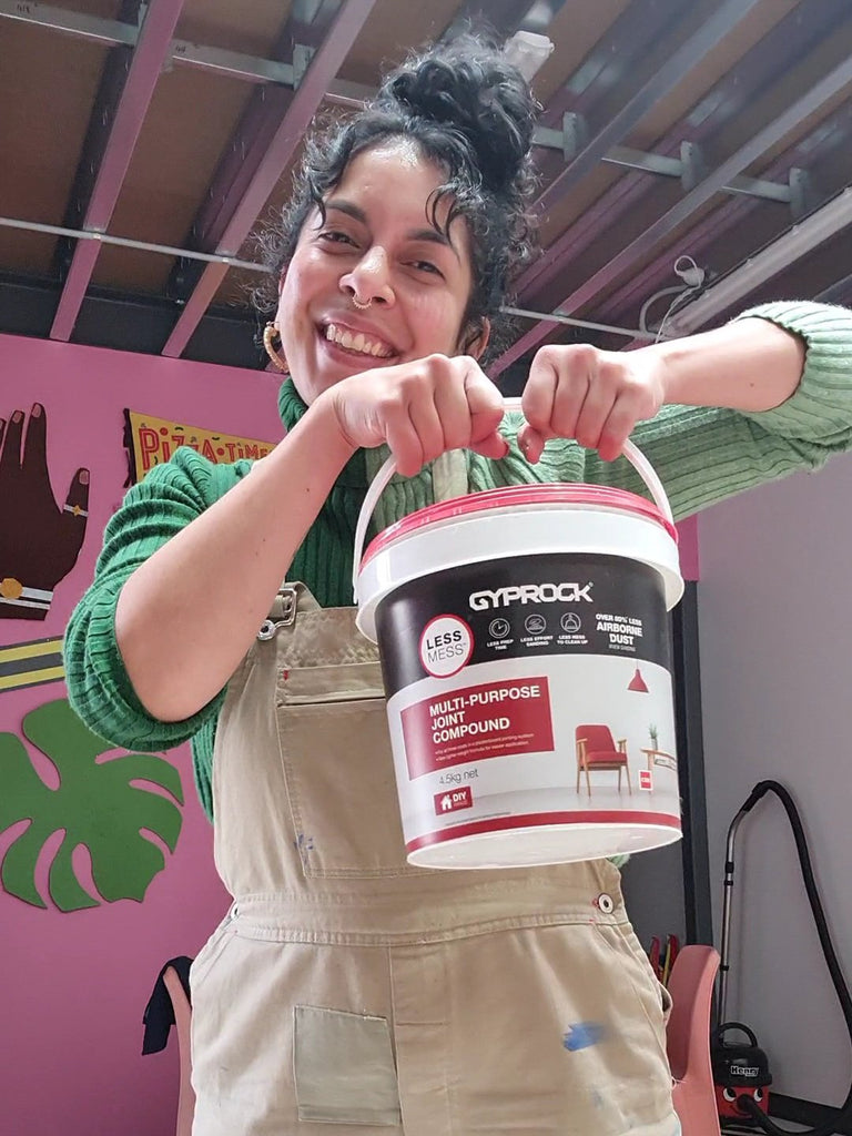 Kitiya, a woman of colour, wearing a green sweater and brown overalls, stands in her colourful art studio holding a tub of multi-purpose joint compound for making papier mache props