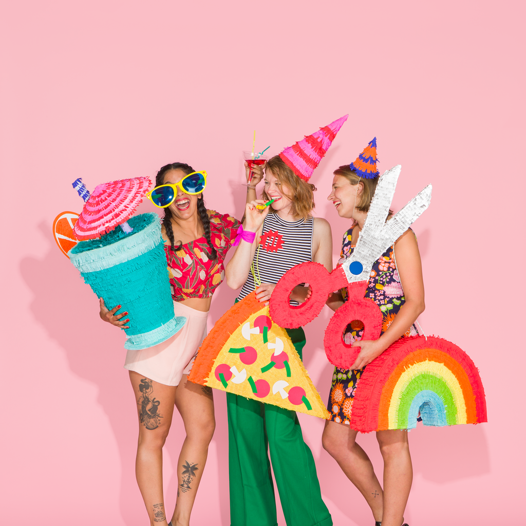 3 women in party outfits hold a variety of colourful and festive pinatas against a pink background