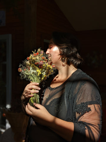 Cinqala, an Indigenous and Romani person with chin-length dark hair, stands partially in shadows in front of a window. They hold a bouquet of yellow, red, and white dried flowers to their face, and are smiling, with their eyes downcast.