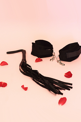 A black flogger and a pair of black cuffs sit atop a pink background, along with a fe red roses.